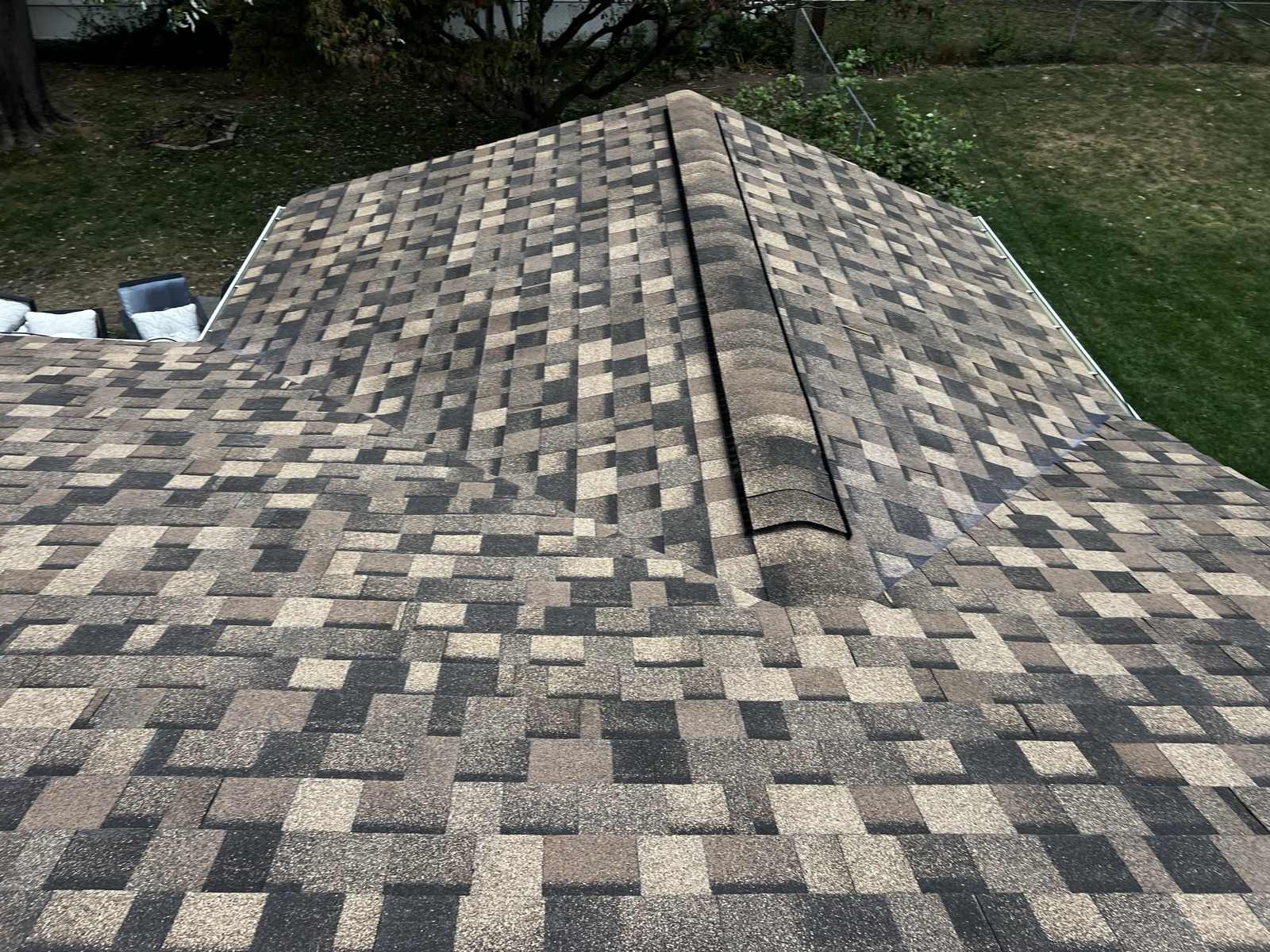 The hip of a newly laid brown, tan and charcoal multi tone architectural shingle roof, capped with ridge cap shingles running up the hip over a dark ridge vent strip, with the two roof planes falling away either side and a mown lawn, a chain link fence and outdoor cushioned furniture below