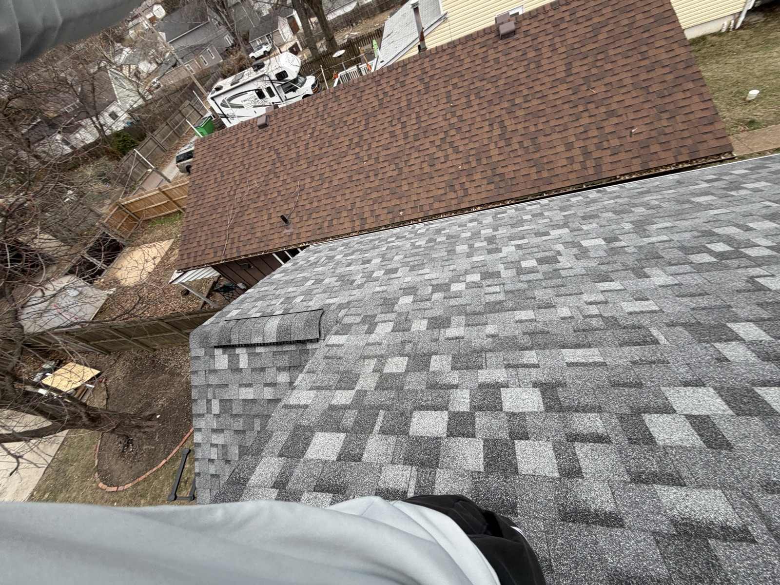 A newly laid grey and charcoal architectural shingle roof with a ridge vent, set directly against the old brown shingle roof of the adjoining garage across the top of the frame, with a back yard, a wooden privacy fence, a white recreational vehicle in the alley beyond and the photographer's knee at the bottom edge