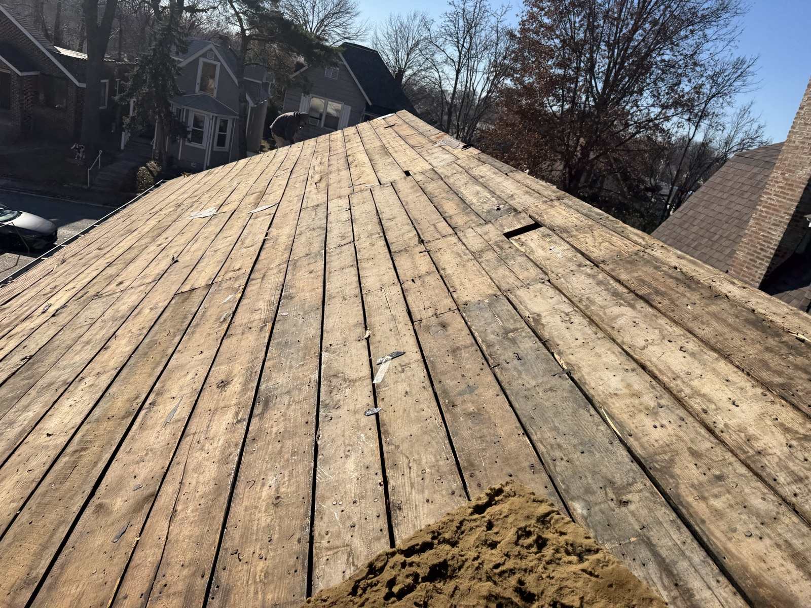 A large roof plane stripped back to bare wooden plank decking, the boards weathered and studded with old nails with scraps of felt caught between them and a pile of loose granules at the bottom right, with a figure bent over at ground level beyond the roof beside a neighbouring grey house, and older brick and frame houses under a clear blue winter sky beyond