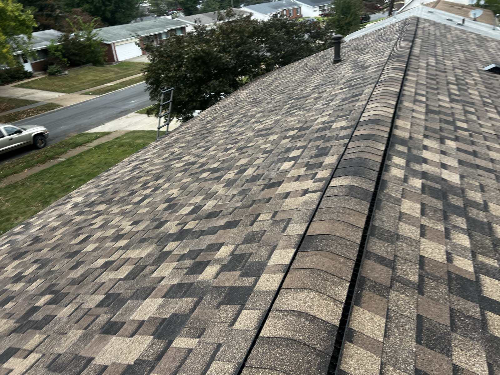 The capped ridge of a newly laid brown, tan and charcoal multi tone architectural shingle roof running diagonally through the frame with a dark ridge vent beneath the cap and a small metal flue near the far end, an aluminium ladder hooked over the eave at the left, and a suburban street of single storey ranch houses below