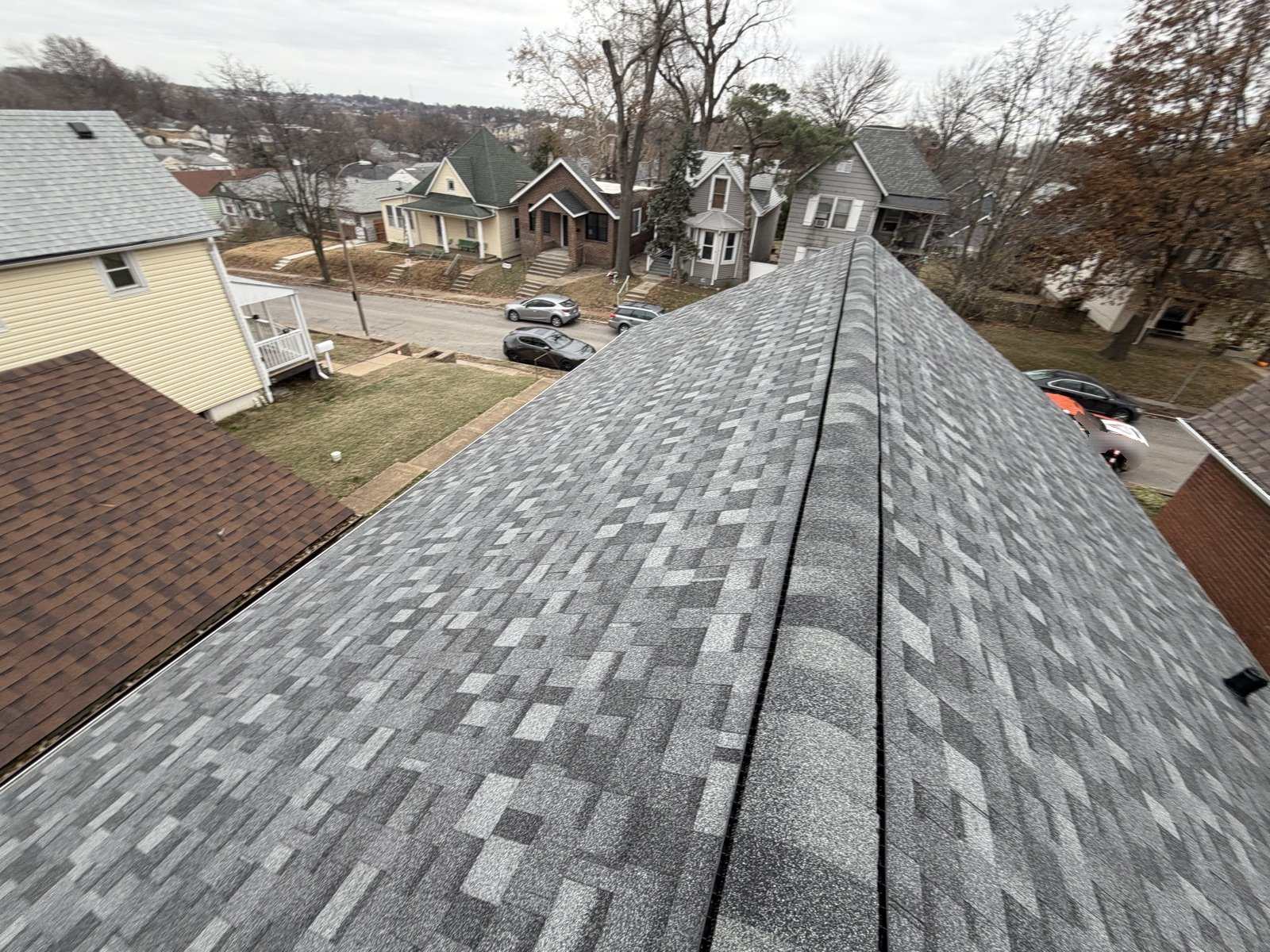 A newly laid grey and charcoal architectural shingle roof photographed along its full ridge, which runs away from the camera with a dark ridge vent along it, looking out over an older St. Louis street of close set houses including a yellow two storey, a green Victorian with a turret and a brick bungalow, with cars parked at the kerb and wooded hills on the horizon