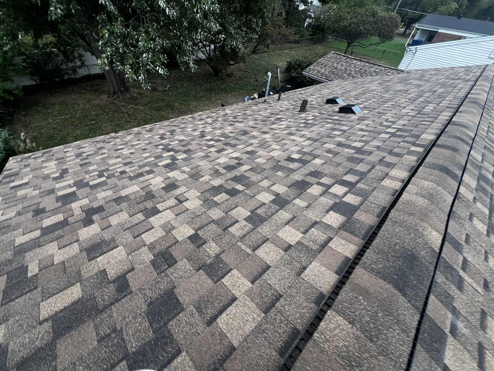 A wide expanse of newly laid brown, tan and charcoal multi tone architectural shingles filling the frame, with a capped ridge and a dark ridge vent along the right side, two black box vents and a metal flue near the far end, and a back yard with a large tree, a chain link fence and a neighbouring white vinyl house below