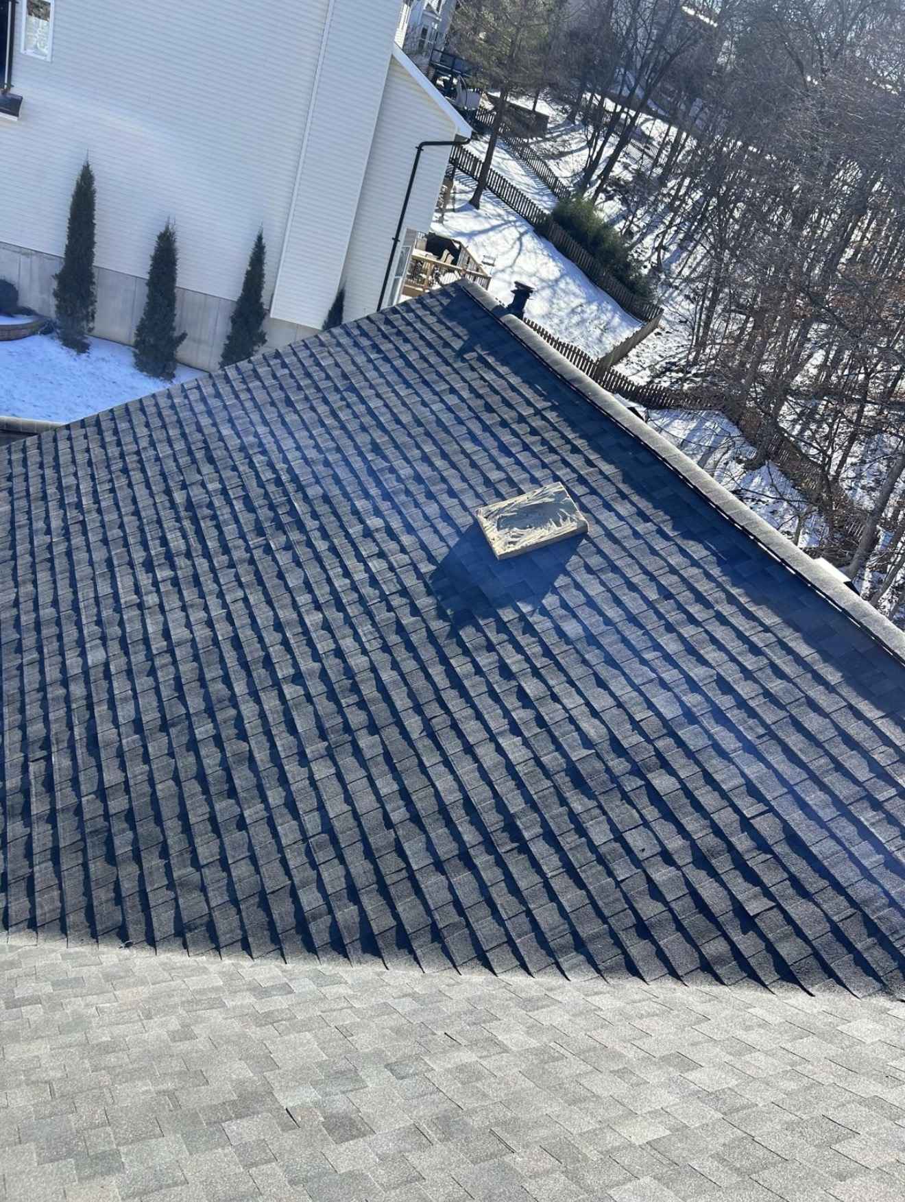 Courses of dark charcoal architectural shingles being laid in a stepped pattern down a steep roof plane, meeting a lighter grey shingle surface across the lower third of the frame, with an offcut of new shingle lying loose on the slope, snow on the ground below and bare winter trees in hard low sun